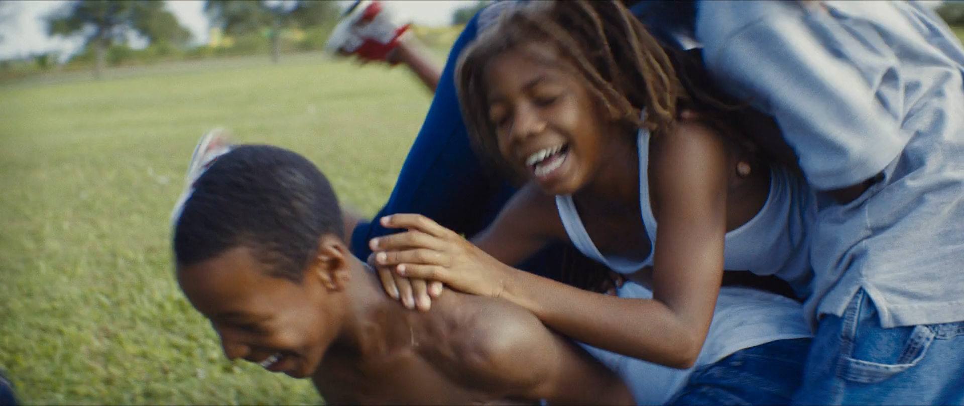 Children wrestle and laugh on green grass, their blue clothing set against warm skin and sunlight.