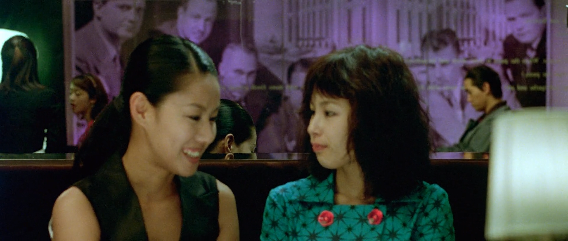 Two women sit together in a dim club washed with purple, green-cyan and small warm accents.