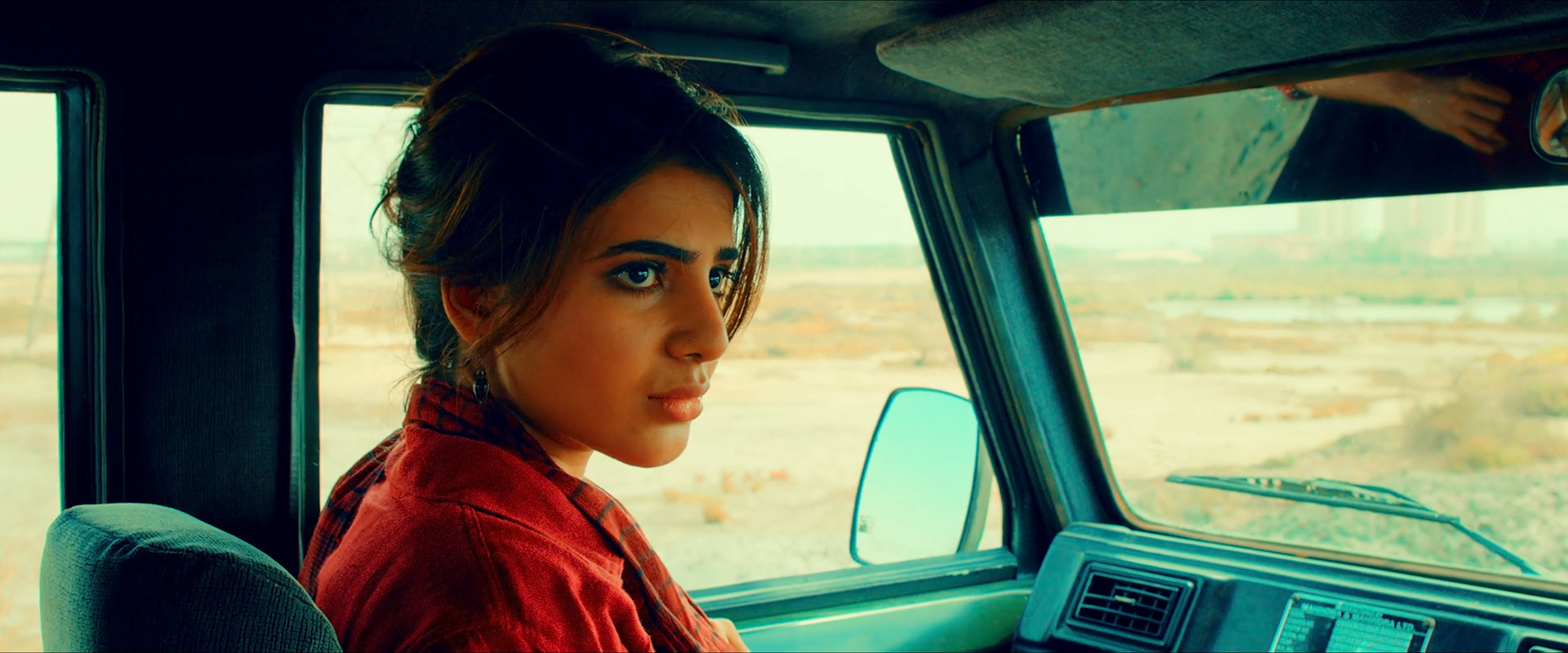 A woman in red sits inside a cyan-blue car interior with warm landscape visible through the window.
