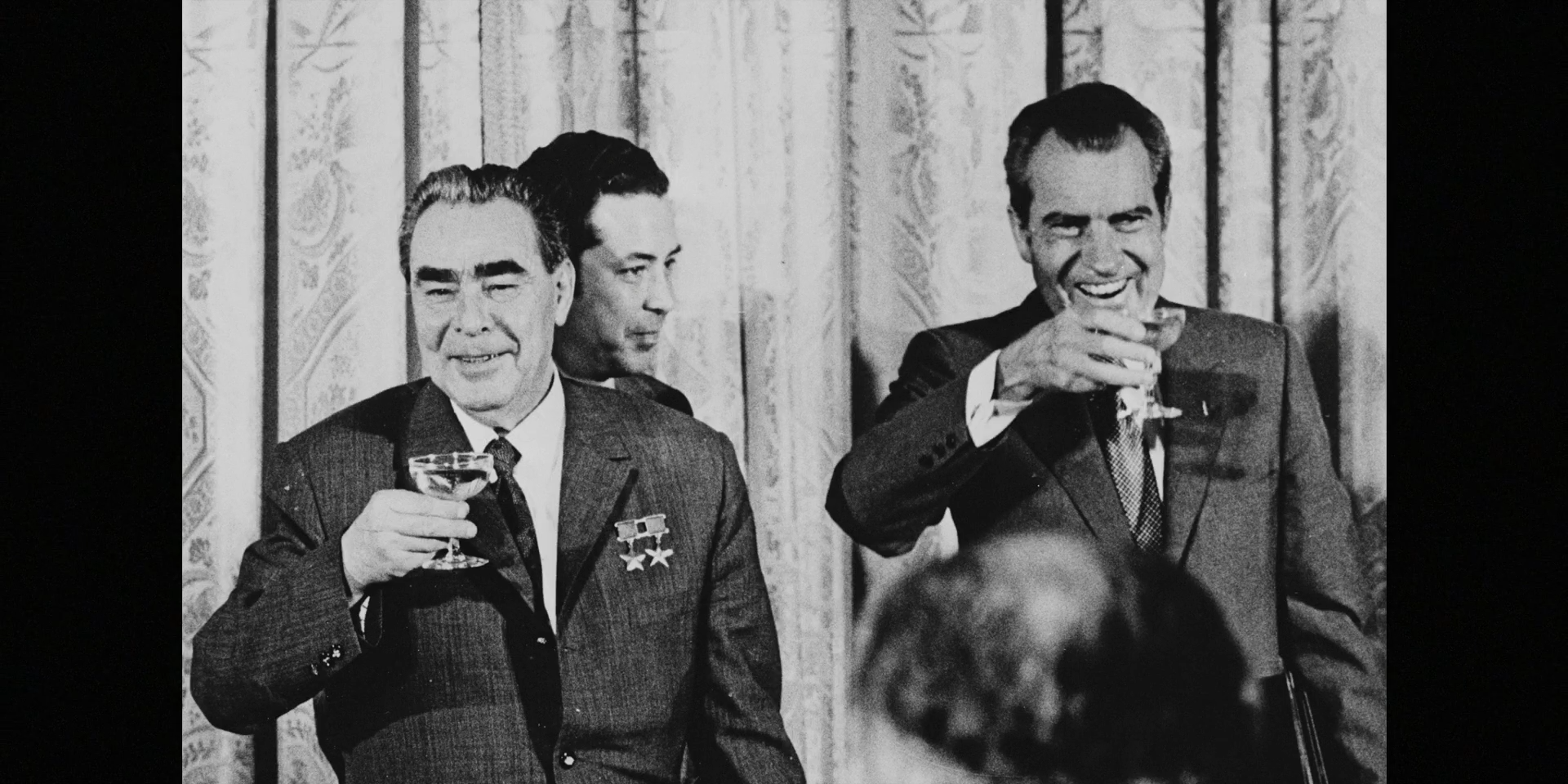 A black-and-white press photograph shows two suited men raising drinks before a patterned curtain.