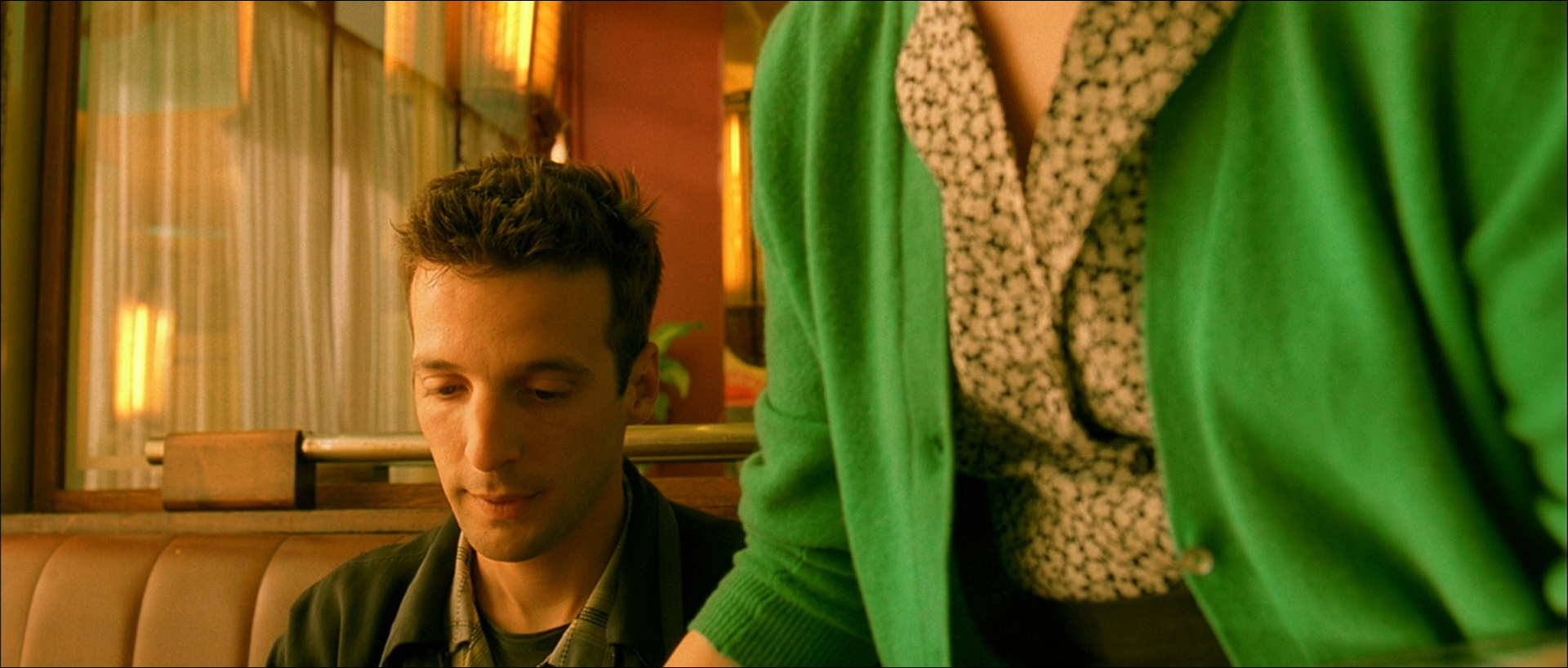 A man sits at a warmly lit diner table while a vivid green jacket fills the foreground beside him.
