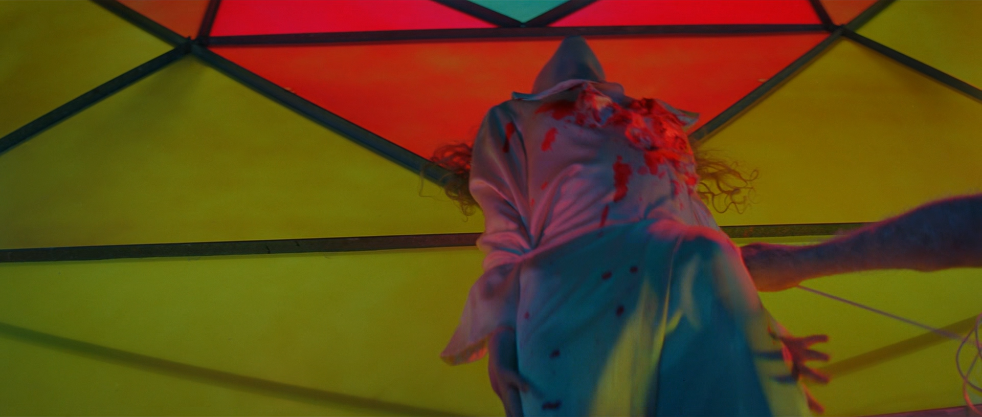 A figure reaches upward beneath a geometric canopy of red, yellow and green translucent panels.