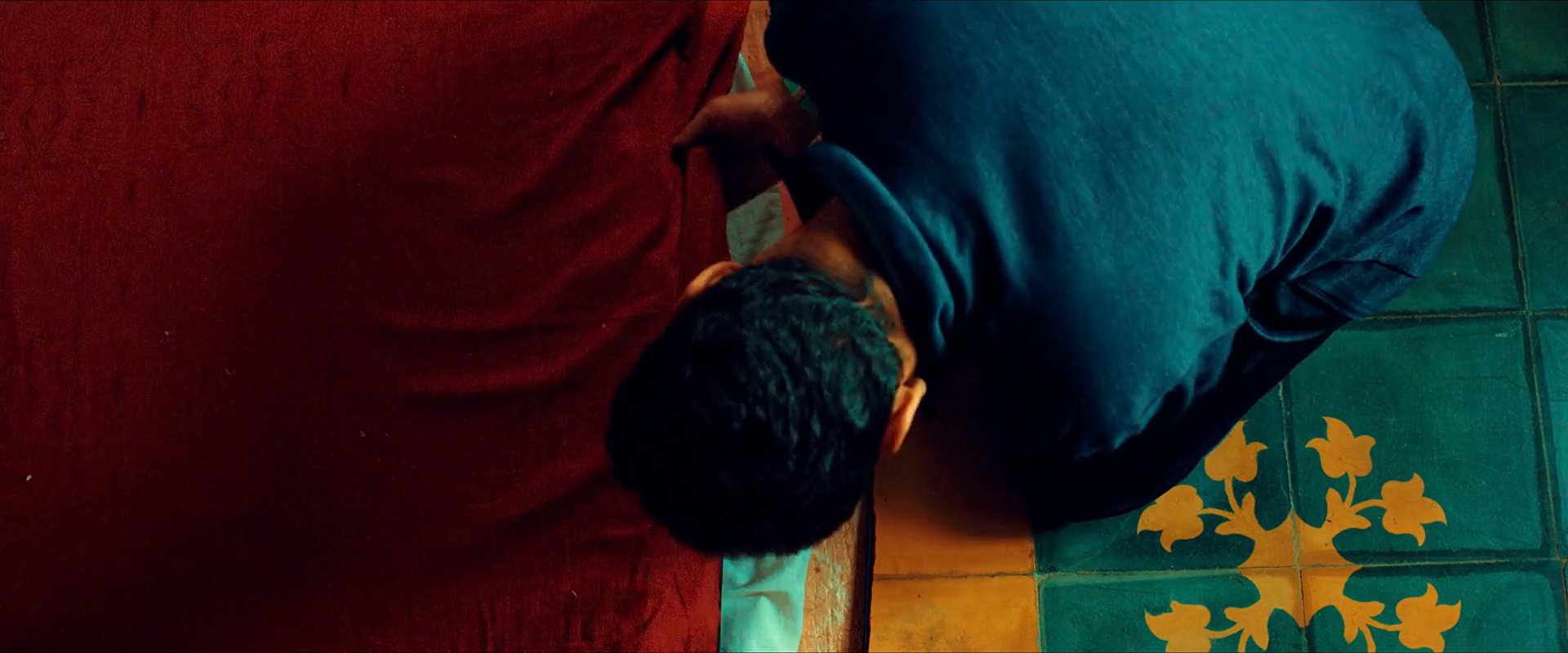 A man curls on a floor divided into saturated red, teal-blue and mustard-yellow areas.