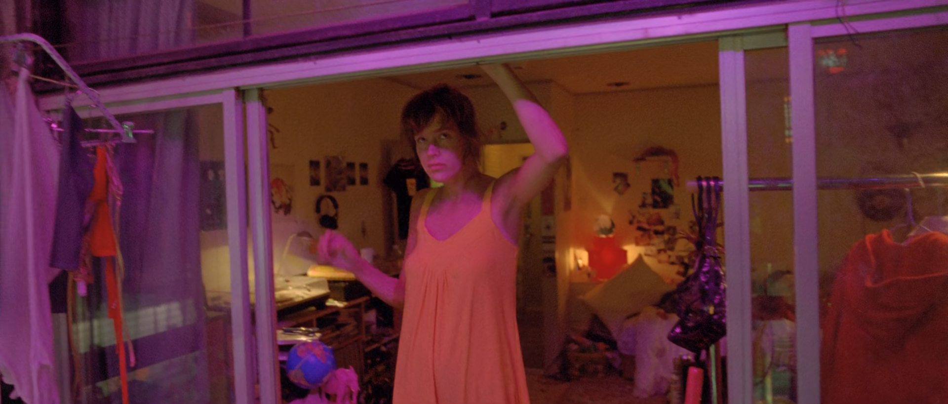 A woman in a pale slip stands in a magenta and violet-lit room divided by glass panels.