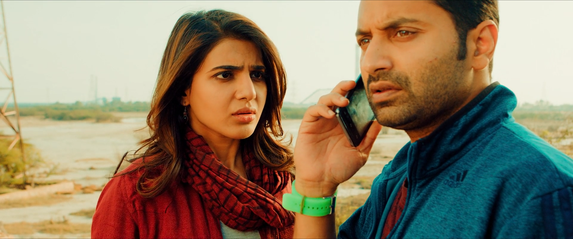 A woman in red stands beside a man in teal on a phone, against dusty yellow-green surroundings.
