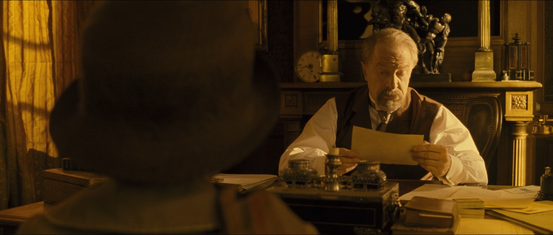 An older man in a waistcoat reads a sheet of paper at a desk in an amber-lit office, seen past a dark out-of-focus figure in a hat.