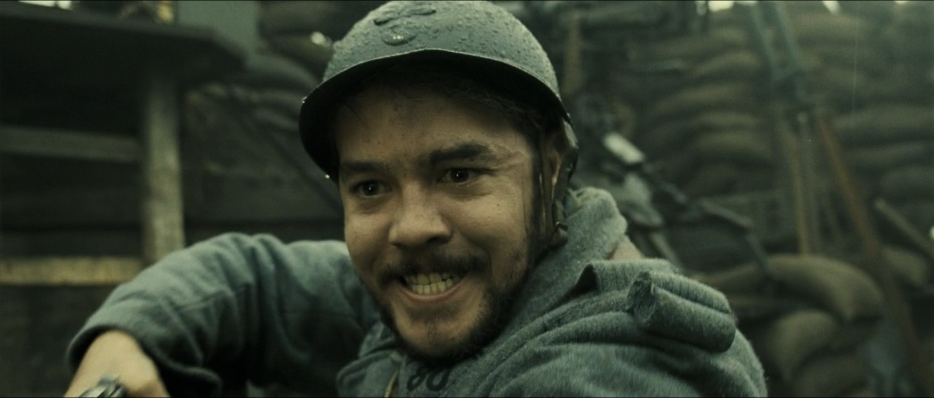 A grinning helmeted soldier leans out of a wet trench, his greatcoat, the sandbags and the timber behind him all neighbouring olive and blue-grey tones.