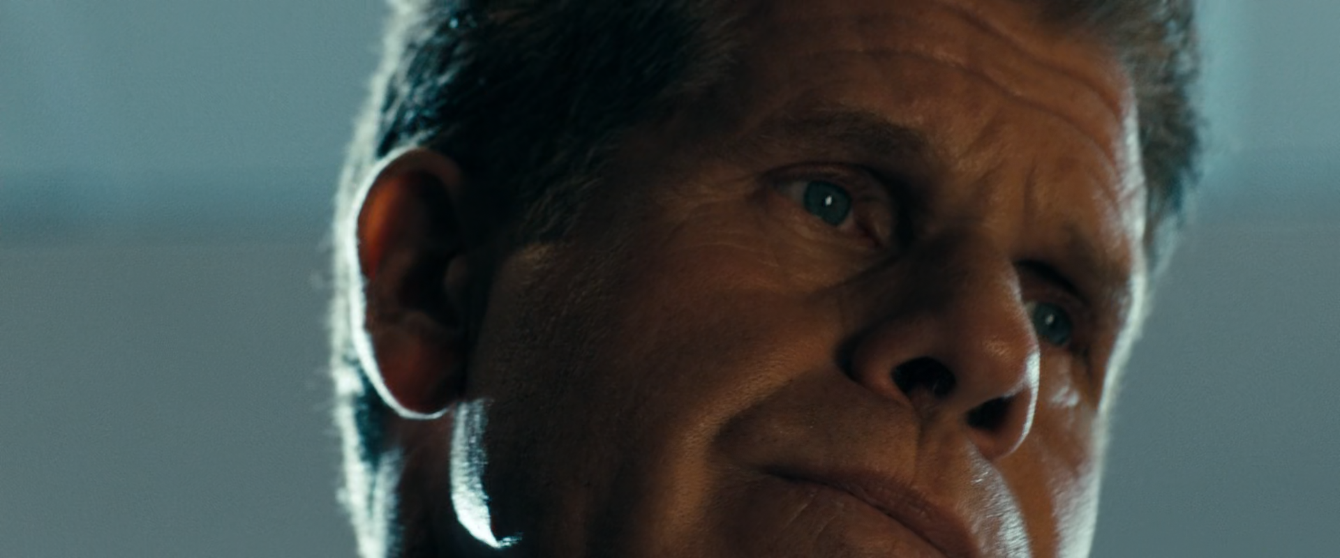 A tight close-up of an older man's warmly lit face turned upward against a cool blue-grey wall.