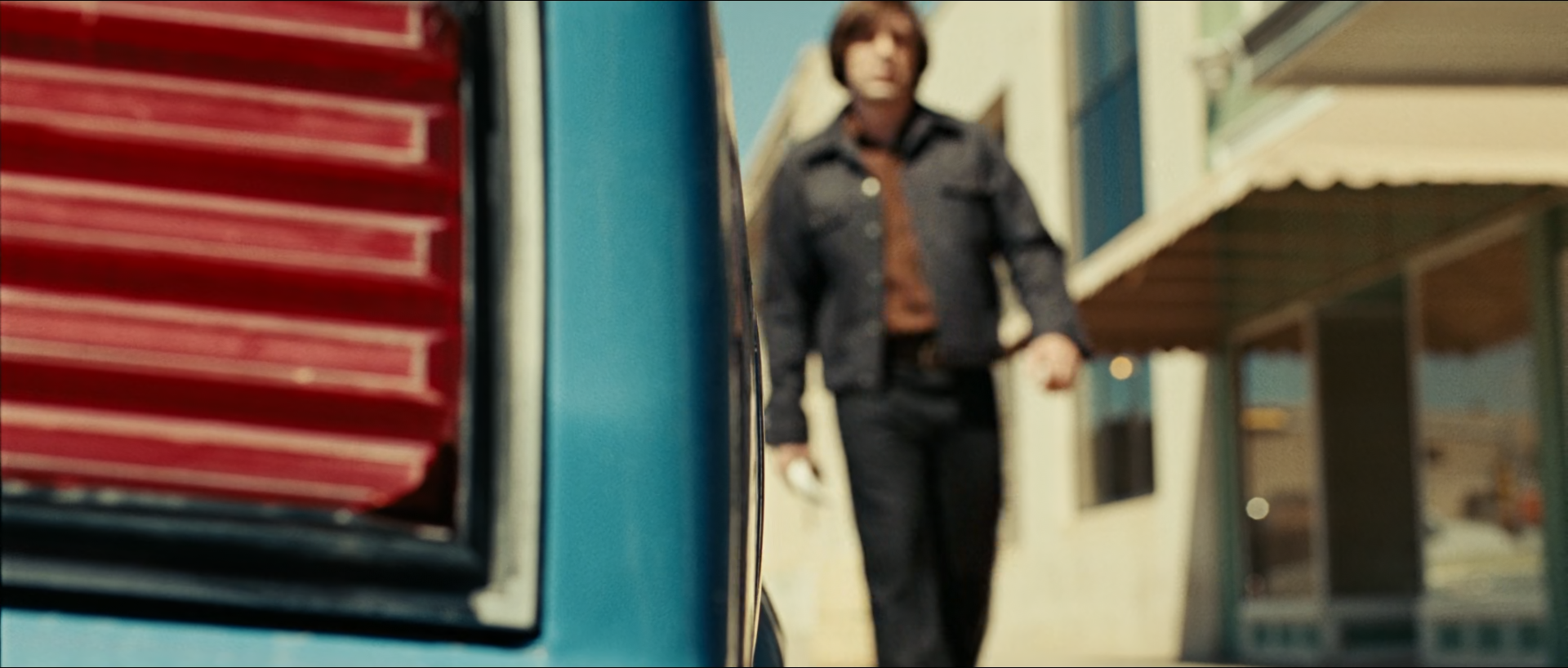 A man in a denim jacket and rust shirt walks past a motel, seen past the red tail light and teal bodywork of a truck filling the left foreground.