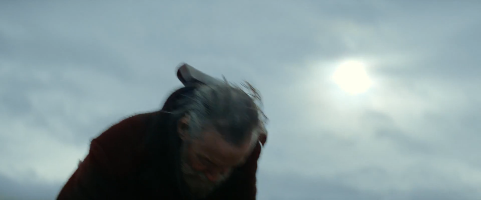 An overcast sky with the sun a soft disc behind cloud, and a man's grey hair below it.