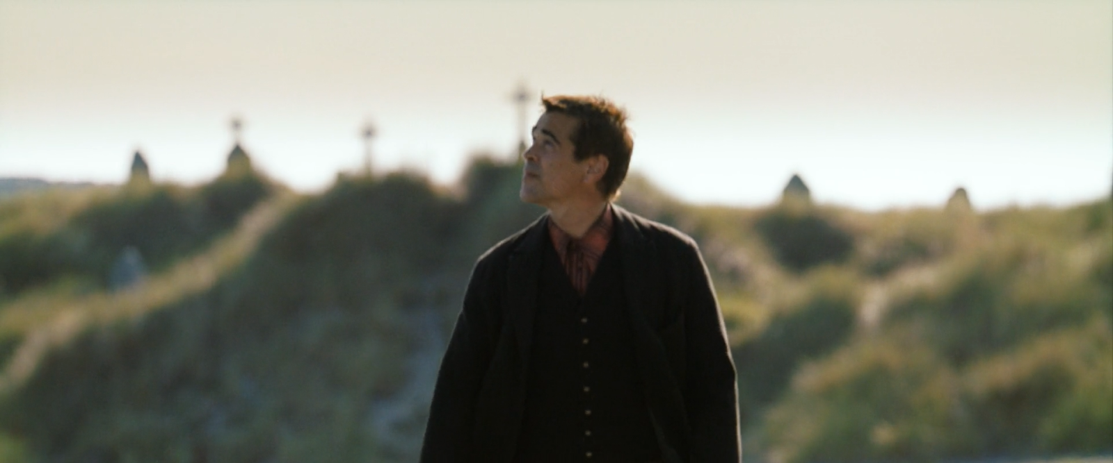 A man in a dark coat against a pale even coastal sky above a dune.