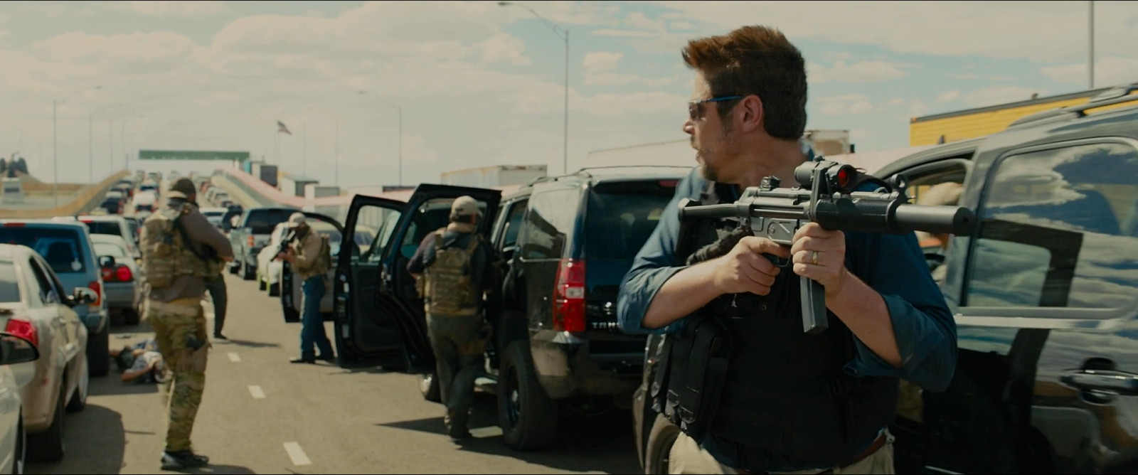 A noon border crossing, cars stopped in lanes, an armed man in the foreground, bright hazy sky above.