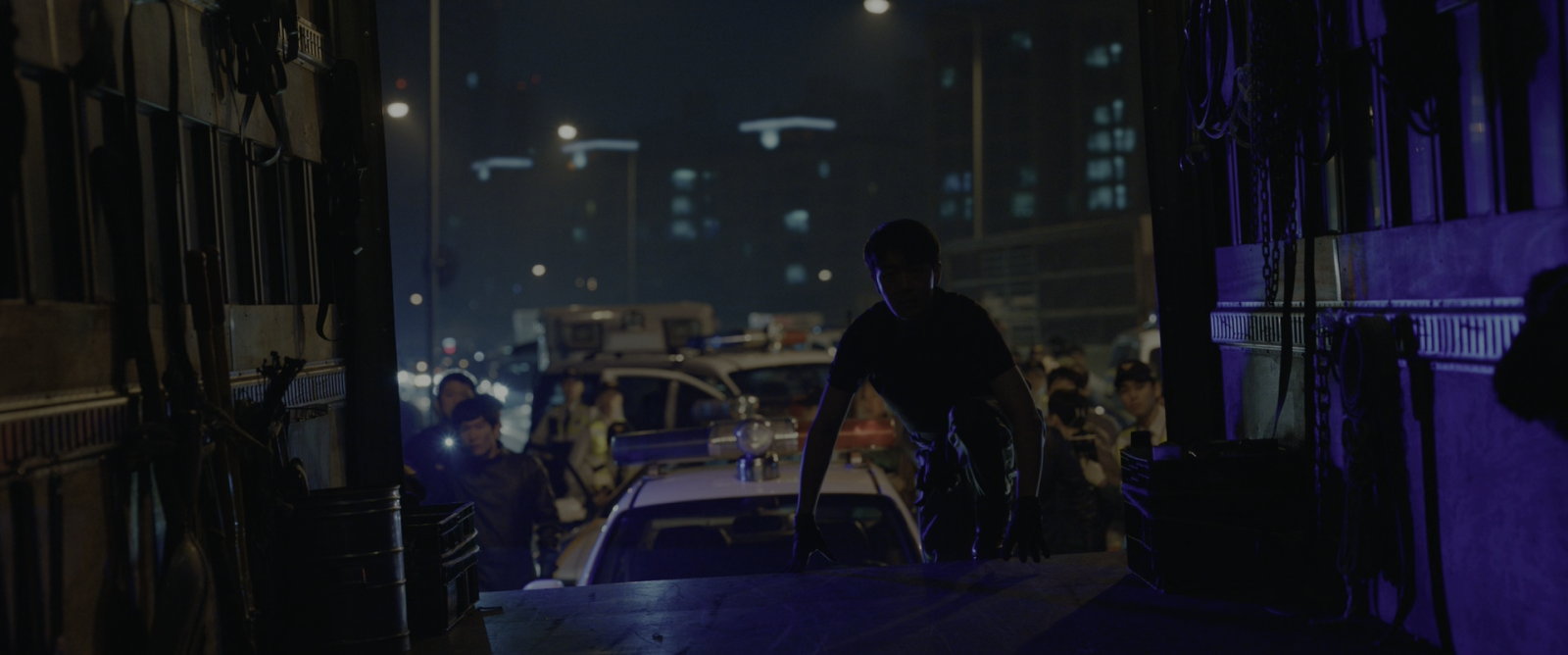 A night street seen from inside a truck, police cars, a crowd and many small practical lights.