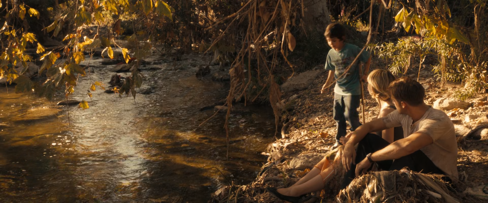 Three people sitting by a sunlit creek under overhanging foliage, the shadows falling in small scattered patches.