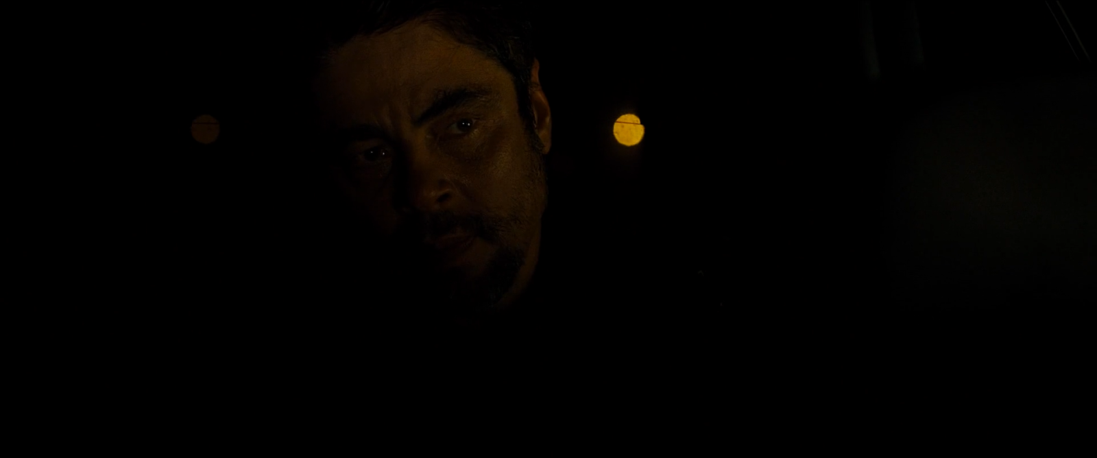 A man's face lit faintly from one side in a car at night, most of the frame at absolute black with two out-of-focus lights behind him.