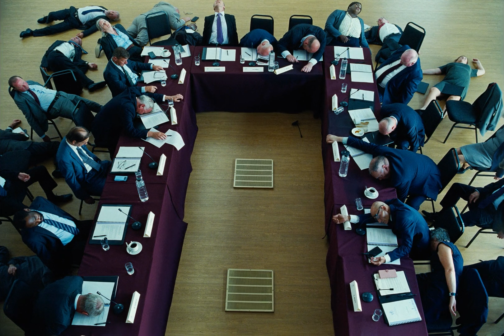 Overhead view of a U-shaped conference table in burgundy cloth on a pale yellow wooden floor, ringed by people in dark blue suits slumped over it and on the ground.