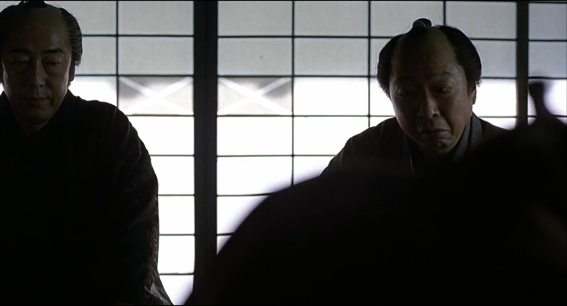Shot 0124 from Zatoichi. Backlit shoji turn white-gray, leaving faces and robes as heavy silhouettes; the foreground blur compresses the image further.