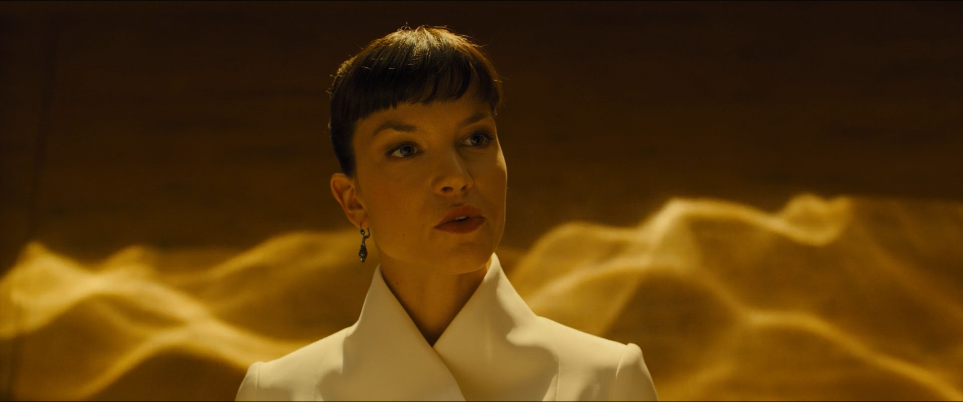 Shot 0326 from Blade Runner 2049 at 01:30:53:12. Reflected water-light crosses the wall in bands of gold, and the white jacket and the face take the same warm cast, so the figure separates by brightness rather than by hue.