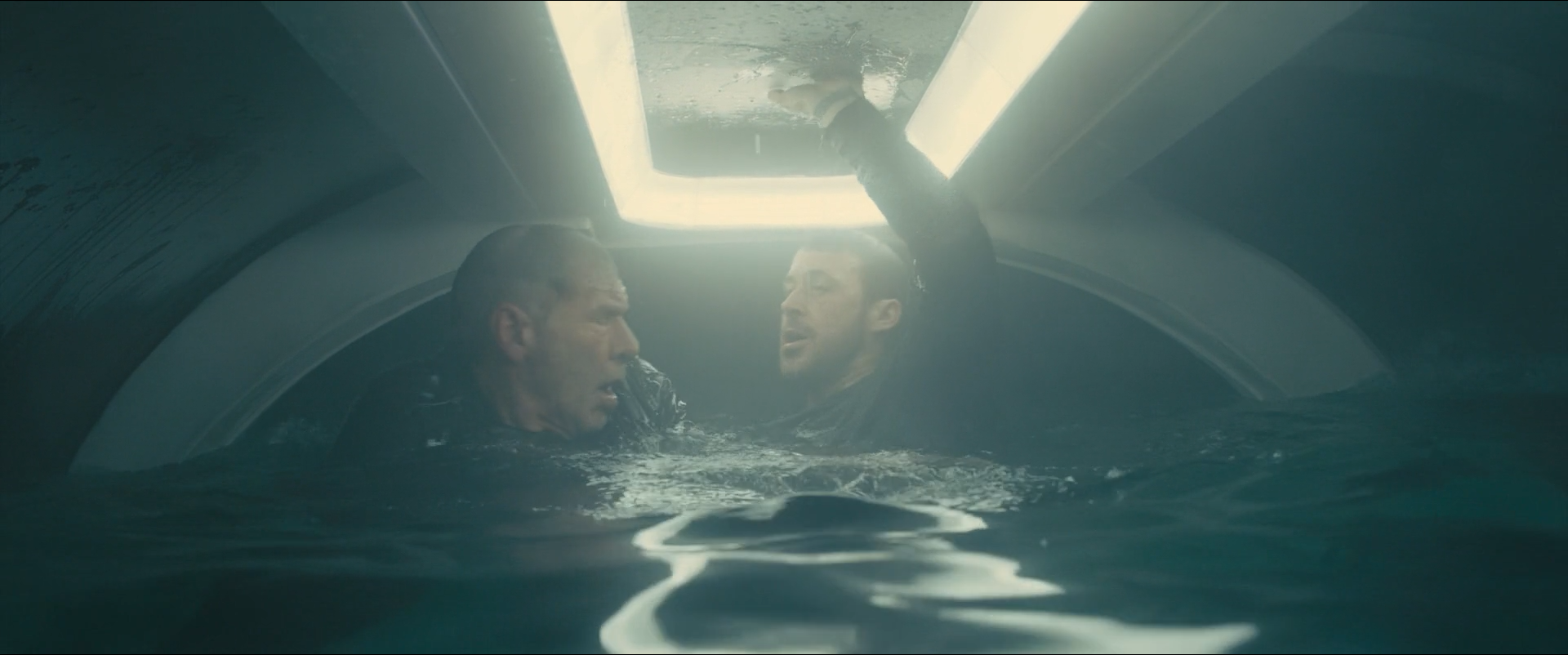 Shot 1863 from Blade Runner 2049 at 03:25:40:02. A single ceiling panel lights the water and both faces from above; the cabin walls and the surface below stay green-black, so the image is built from one reflected highlight.
