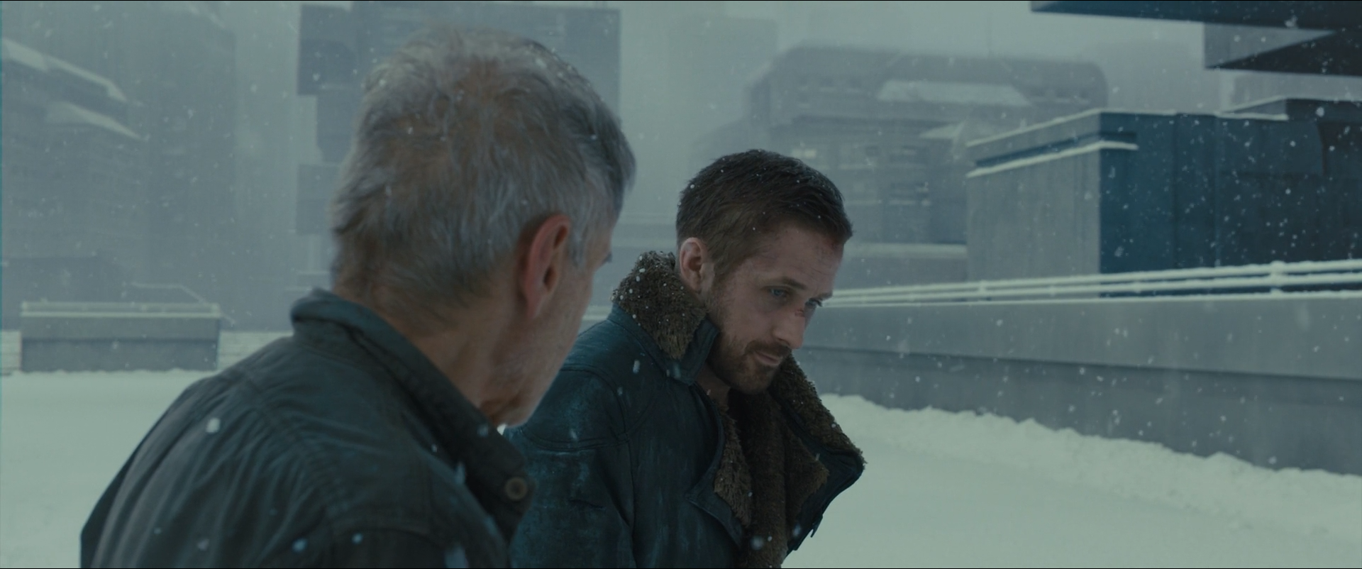 Shot 1892 from Blade Runner 2049 at 03:29:16:01. Snow and pale concrete hold the background at an even light grey; the two dark coats and the fur collar are the only dense masses, and the flakes give the air its texture.