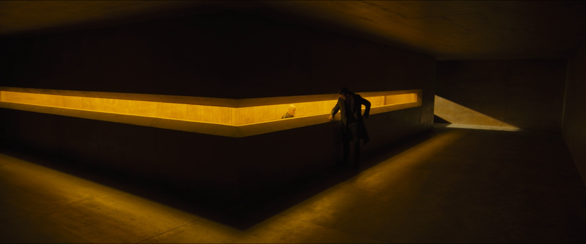 Shot 0316 from Blade Runner 2049 at 01:29:38:12. One continuous light trough draws a shallow V across dark concrete; everything above and below falls to near-black, and the leaning figure is placed exactly at the join.