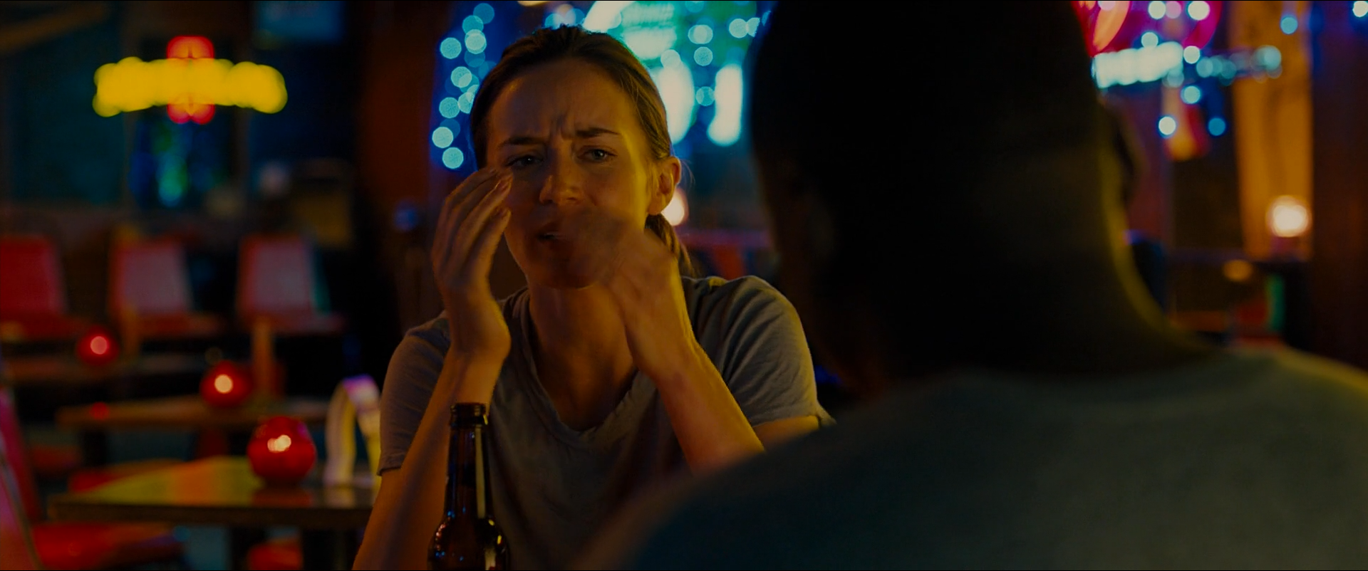 Shot 0647 from Sicario at 02:05:55:08. Scattered bar signs throw red, blue and yellow points across a dark room; Kate is lit by a low practical while the man opposite is held as a black mass in the foreground.