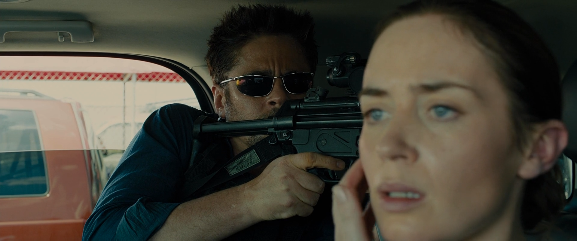 Shot 0376 from Sicario at 01:35:51:15. Shallow focus keeps Kate sharp at right while the barrel and the man behind it stay soft; the red vehicle beyond the window is the frame's only strong colour.