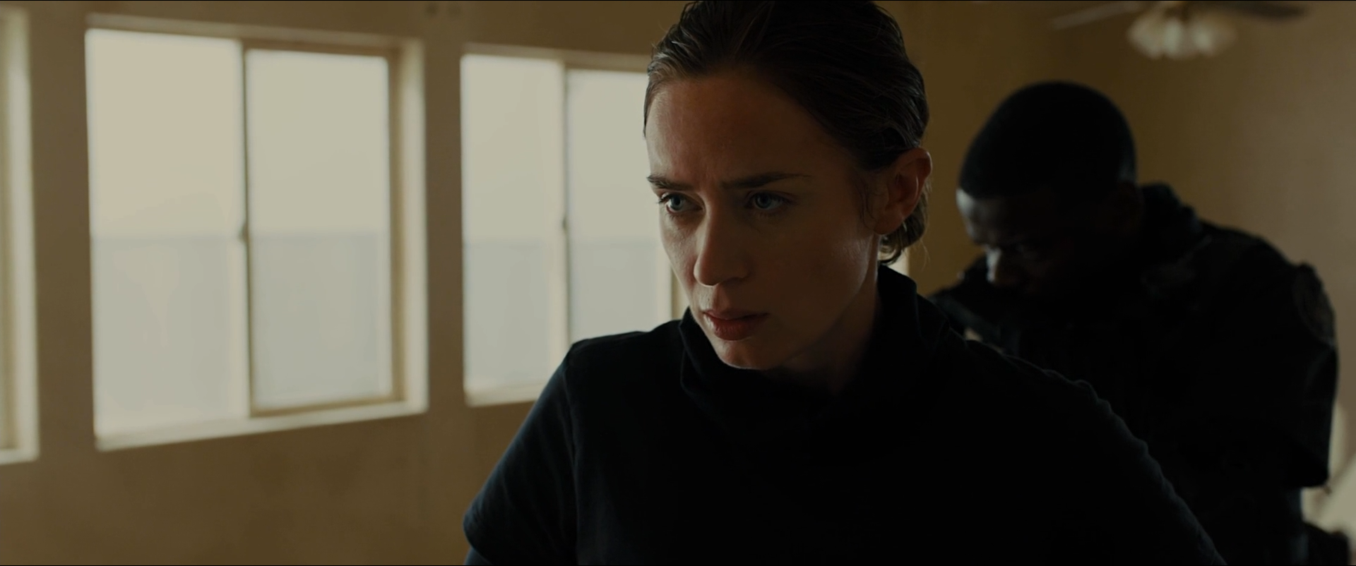 Shot 0062 from Sicario at 01:05:05:09. Two window panels clip to white behind Kate while the interior falls away to near-black; her face and the shoulder of the figure behind her are the only mid-tones present.