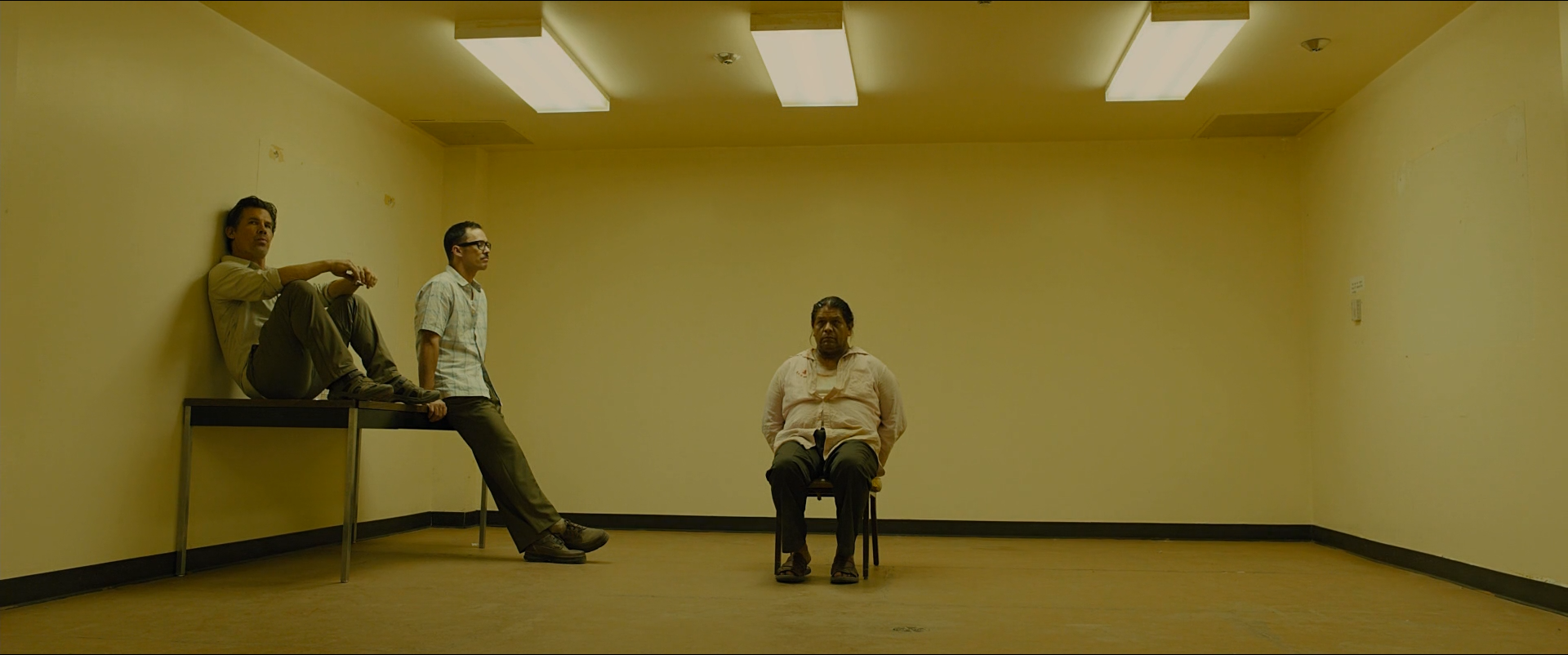 Shot 0454 from Sicario at 01:42:08:00. Fluorescent ceiling panels and yellow walls fill the frame with a single institutional hue; the seated man at centre and the two waiting at left are placed against an otherwise empty box.