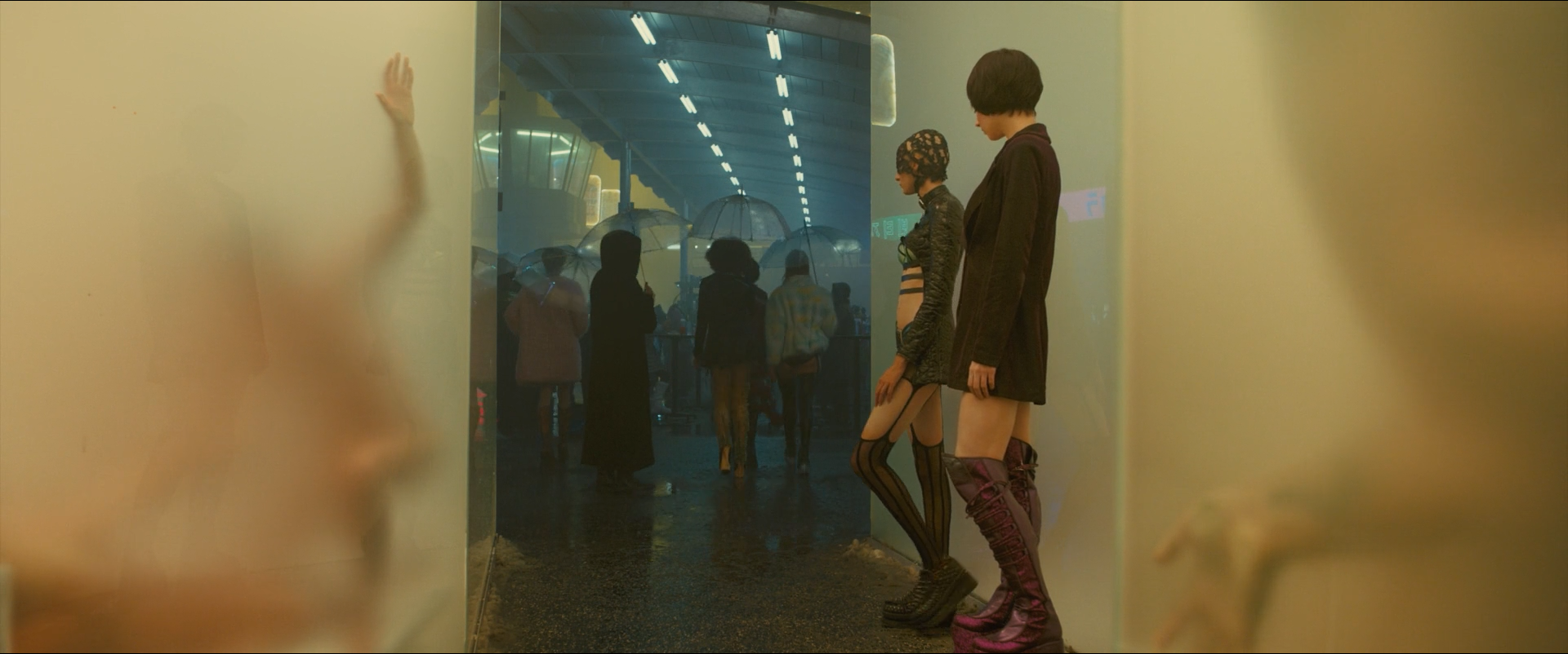 Shot 0434 from Blade Runner 2049 at 01:44:11:04. Frosted panels at both edges carry pressed hands and a blurred face, and a cold, crowded arcade shows through the gap between them; two figures in dark clothing stand on that boundary.