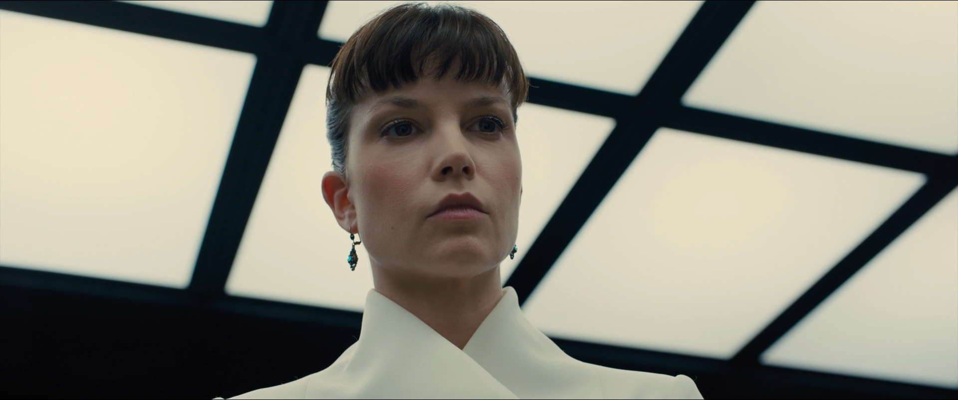 Shot 0359 from Blade Runner 2049 at 01:34:31:08. Lit ceiling panels blow to white and throw a hard graphic lattice behind the head; the pale coat picks up that light while the frame holds almost no colour.