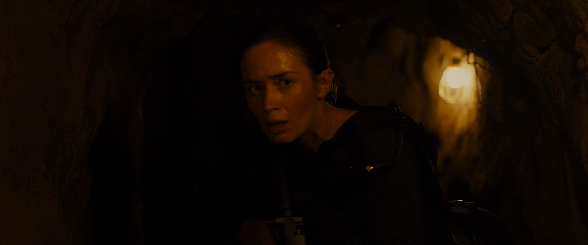 Shot 0931 from Sicario at 02:30:17:16. A single bulb behind her shoulder is all that reaches the frame; the face is modelled by a narrow warm fall-off and the walls disappear entirely.