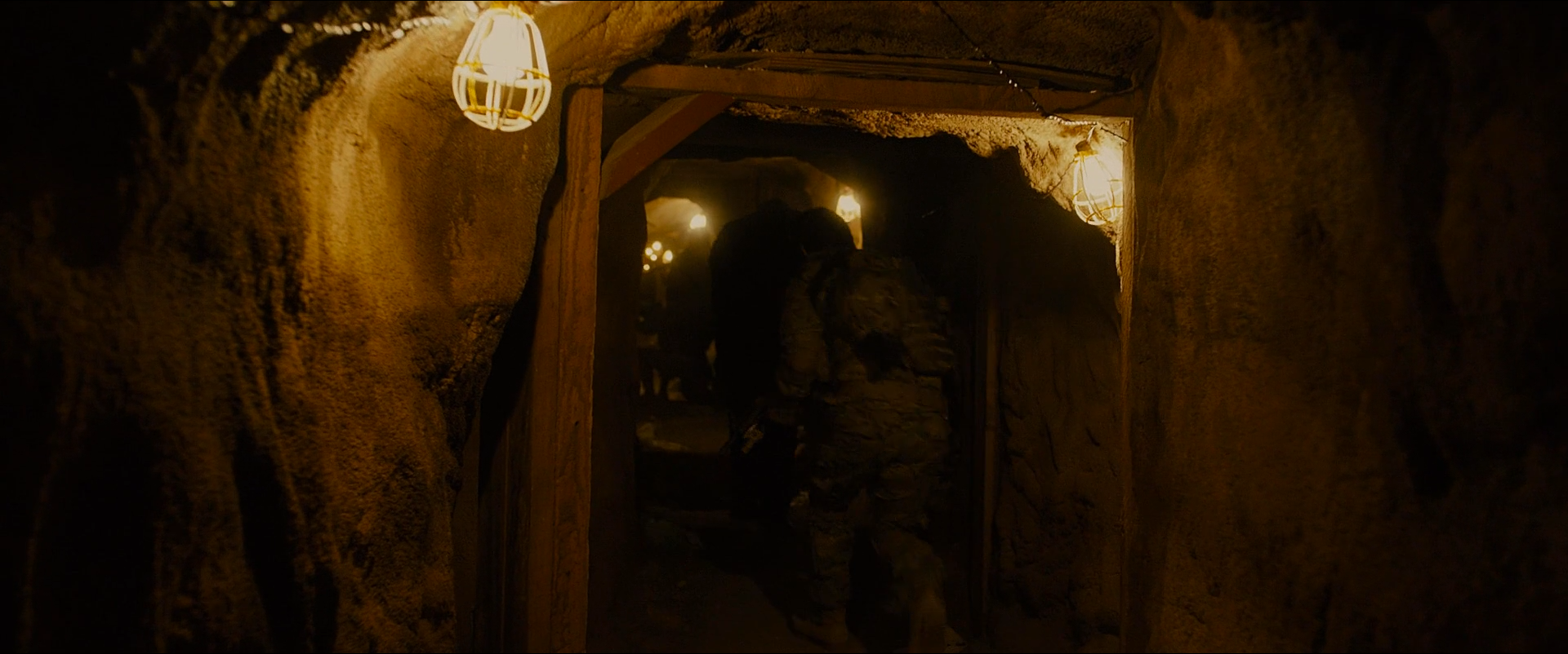 Shot 0887 from Sicario at 02:29:52:04. Two caged bulbs and a receding string of lights are the only sources; the earth walls take their amber directly, and the figures in the doorway are almost fully absorbed.