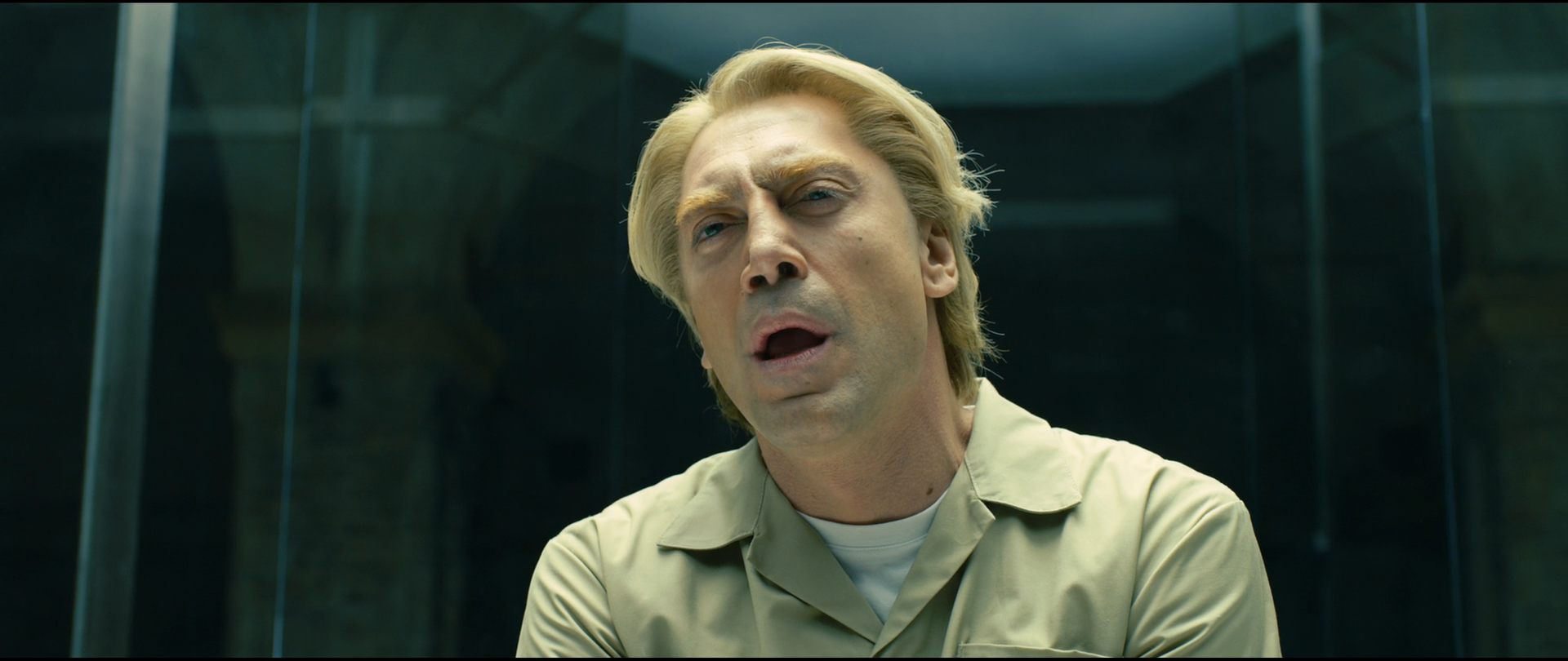 Shot 1389 from Skyfall at 02:23:10:21. The cell walls tint the image a cool green-grey, against which the olive shirt, pale hair and skin are the only warm elements; dark glass mullions frame the head on both sides.