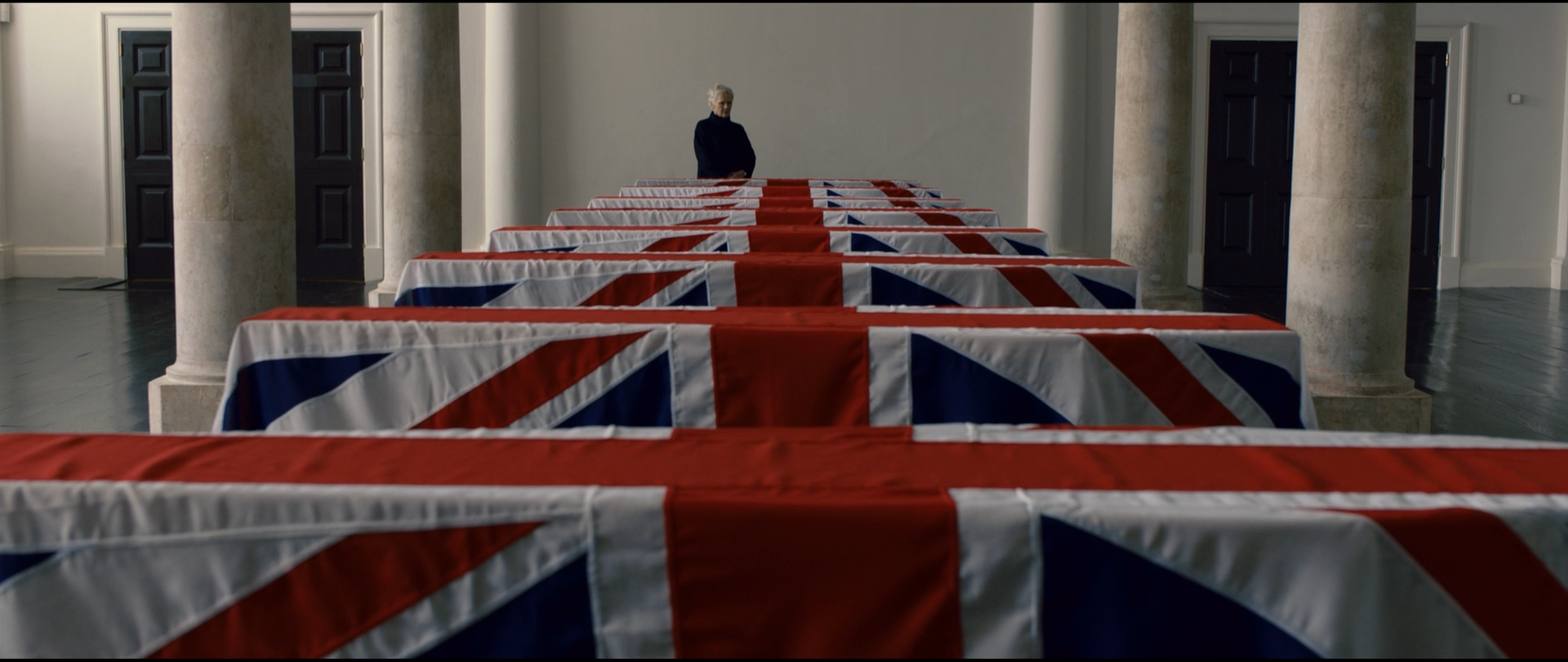 Shot 0613 from Skyfall at 01:24:34:10. Receding rows of Union Jacks drive a red, white and blue perspective into the frame; the stone columns and walls stay near-neutral, and a single dark figure stands at the vanishing point.