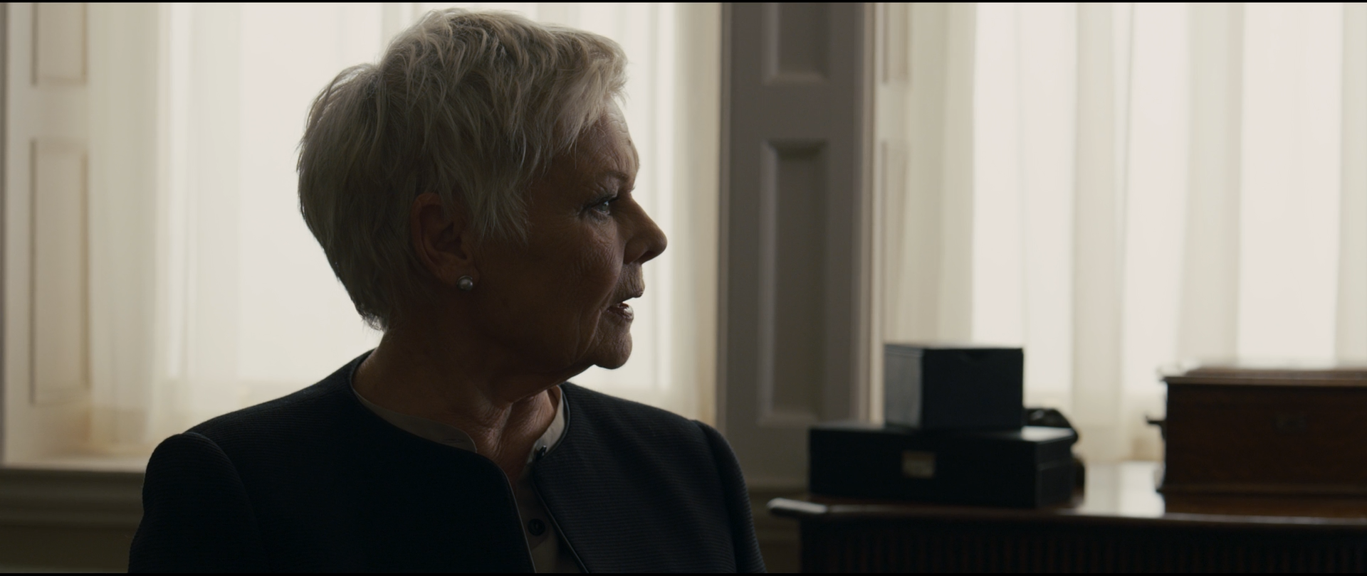 Shot 0529 from Skyfall at 01:18:20:15. Tall curtained windows are allowed to burn out behind M, so her white hair and lit profile sit against blank white while her black jacket merges into the unlit foreground desk.