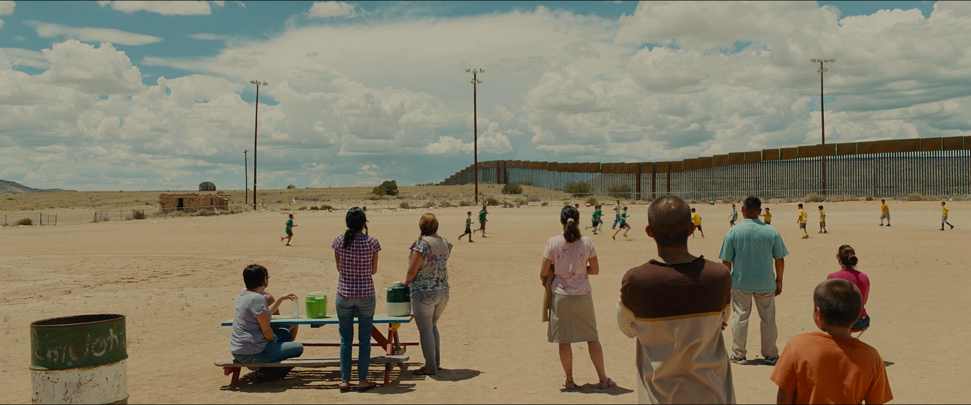 Shot 1202 from Sicario at 02:54:32:08. Bright cloud and pale sand fill the frame; the watching families in the foreground and the border fence across the middle distance divide it into three flat bands.