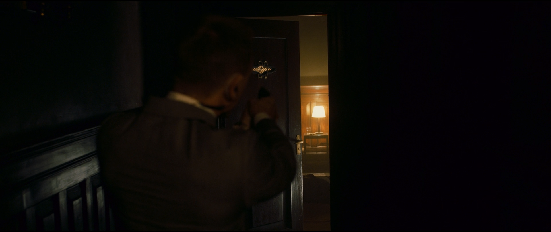 Shot 0002 from Skyfall at 01:01:06:07. Bond's grey shoulder and the small pool of lamplight through the open door carry the whole image; the surrounding wall and banister are held so far down that the frame reads as two lit fragments in black.