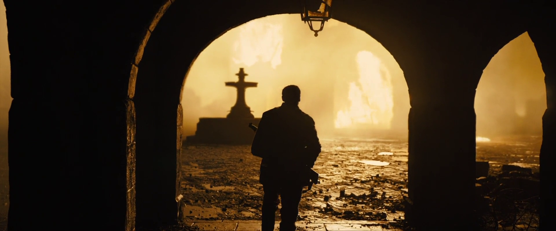Sample 2132 from 1917 at 1:11:06. A black arch frames a square lit only by fire, with a stone cross and a wet street beyond; the man in the opening keeps no detail, and the contrast across the frame is extreme.