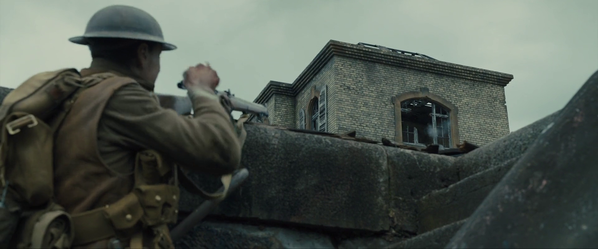 Sample 1952 from 1917 at 1:05:06. A gutted building stands against pale cloud with its windows as black rectangles; the aiming figure in the foreground is dark, and the whole image runs cool.