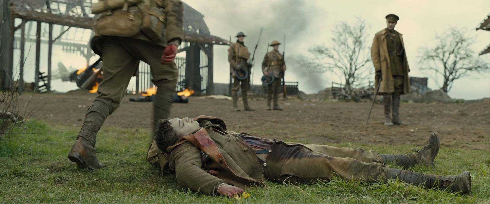Sample 1592 from 1917 at 0:53:05. Green pasture, a burning frame building and standing figures divide the image into three bands; the fallen man in the foreground is the only element at rest.