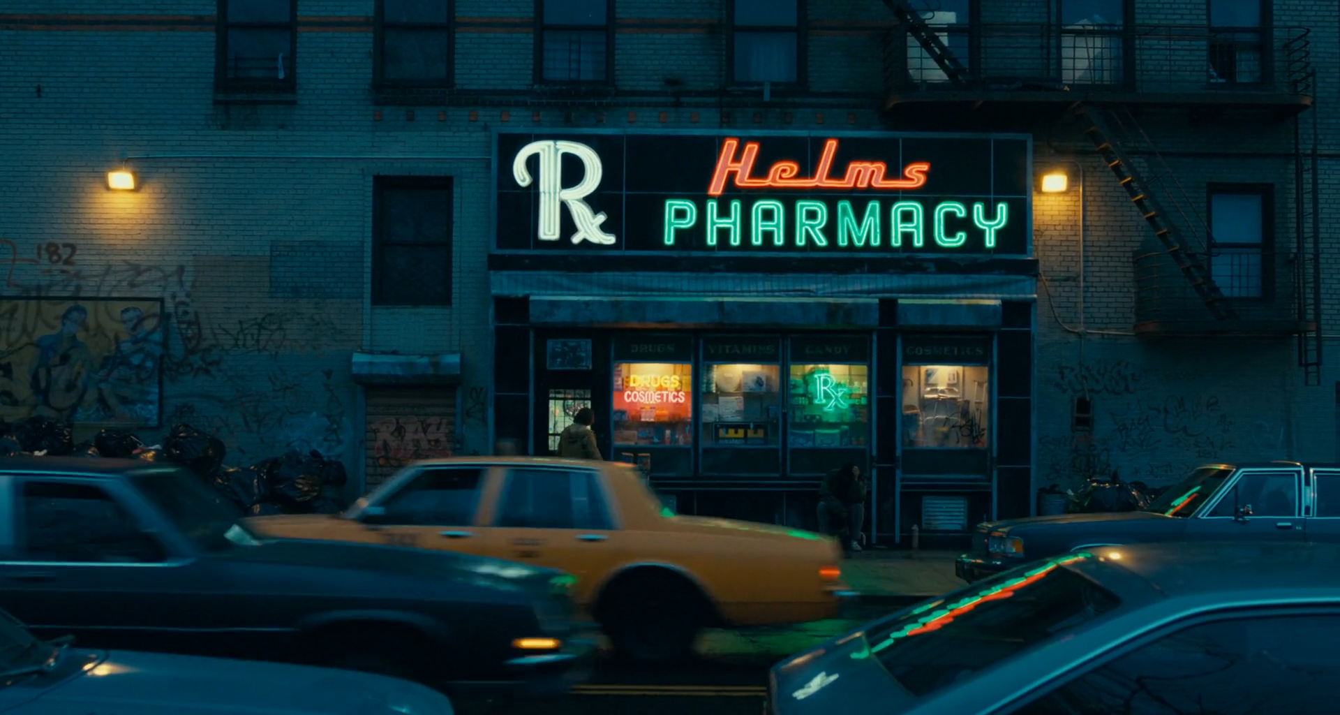 A brick building at dusk with a white, red and green neon pharmacy sign above lit windows, a yellow taxi and dark cars blurred in front.