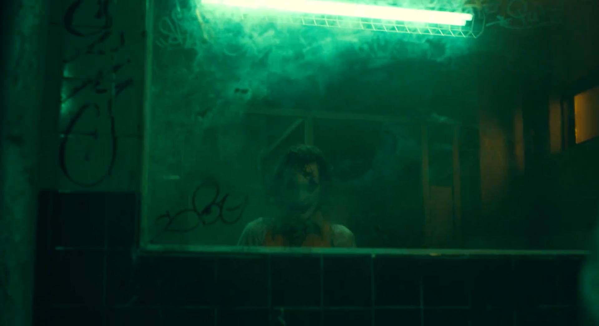 A figure half-seen through a grimy mirror under a green fluorescent tube in a tiled room.