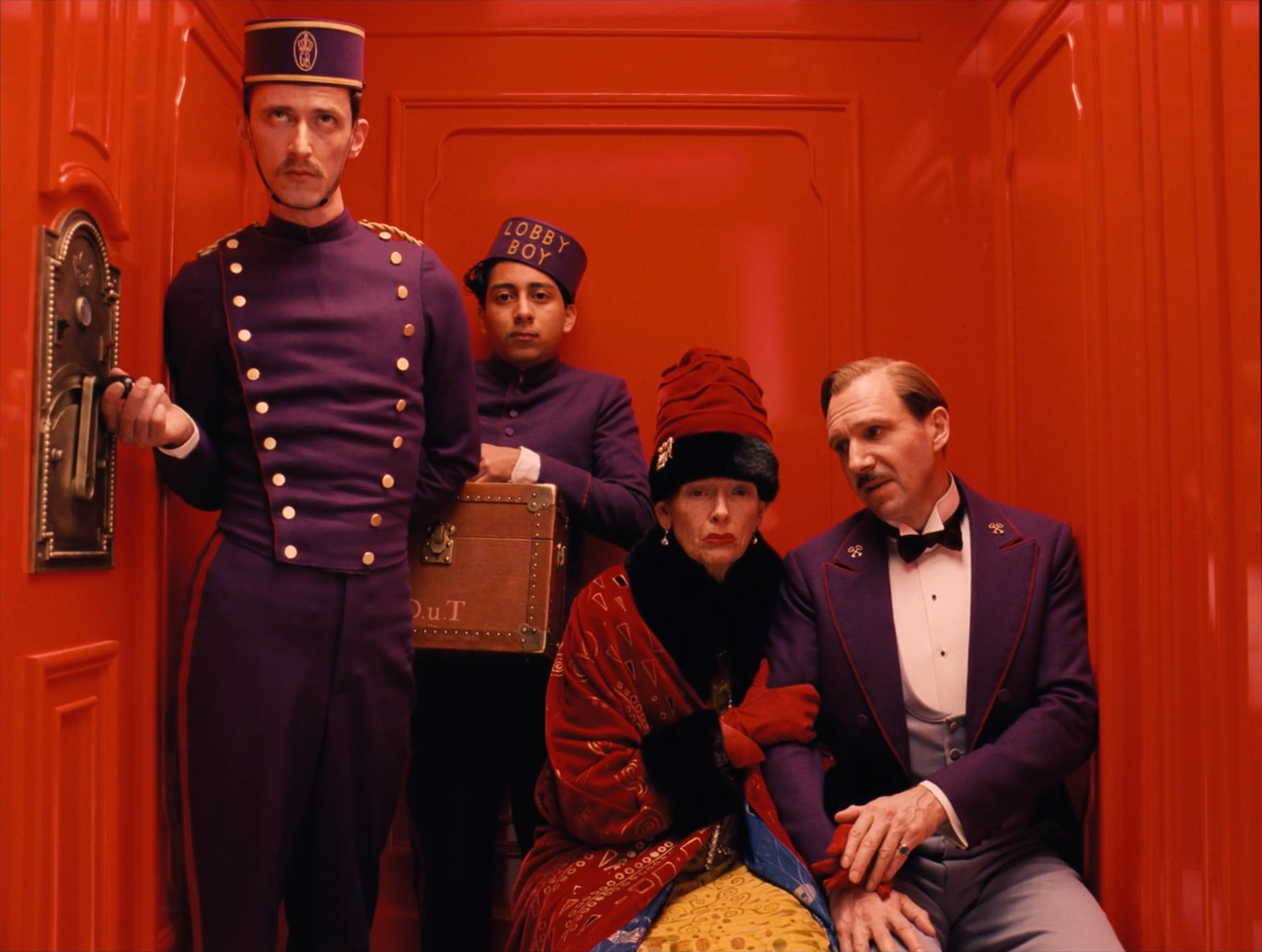 Four people in a red lacquered elevator: an operator and a lobby boy in purple, an elderly woman in a red coat, a concierge in purple.
