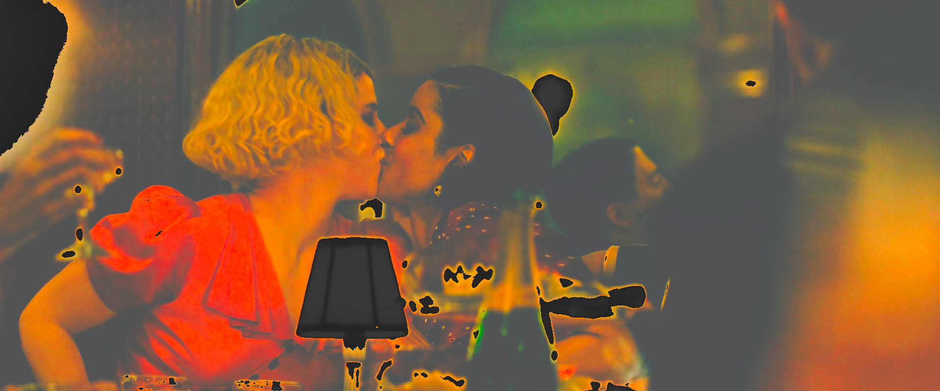 Two women kissing beside a small table lamp in a dark restaurant, the dark lifted to a warm brown-orange.