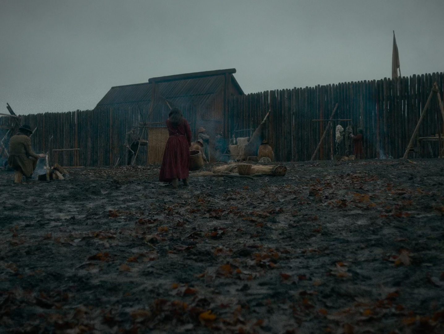 A muddy fort compound with a timber palisade and shingled shed under a flat pale sky; a woman in a dark red skirt stands in the middle ground among smaller figures.