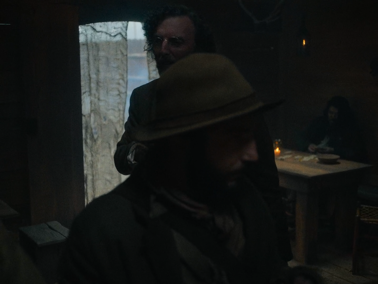 A dark cabin interior seen past a foreground hat brim and shoulder, with a bright curtained doorway at the left, a man in spectacles beyond it, a candle on a table and a woman seated at the right.