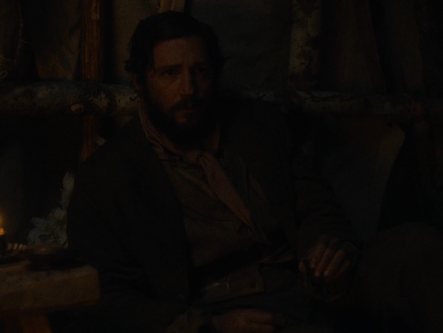 In near-total darkness, a bearded man in a heavy coat leans back against wooden poles and hanging cloth, his face and hands faintly lit by an unseen fire to the left.