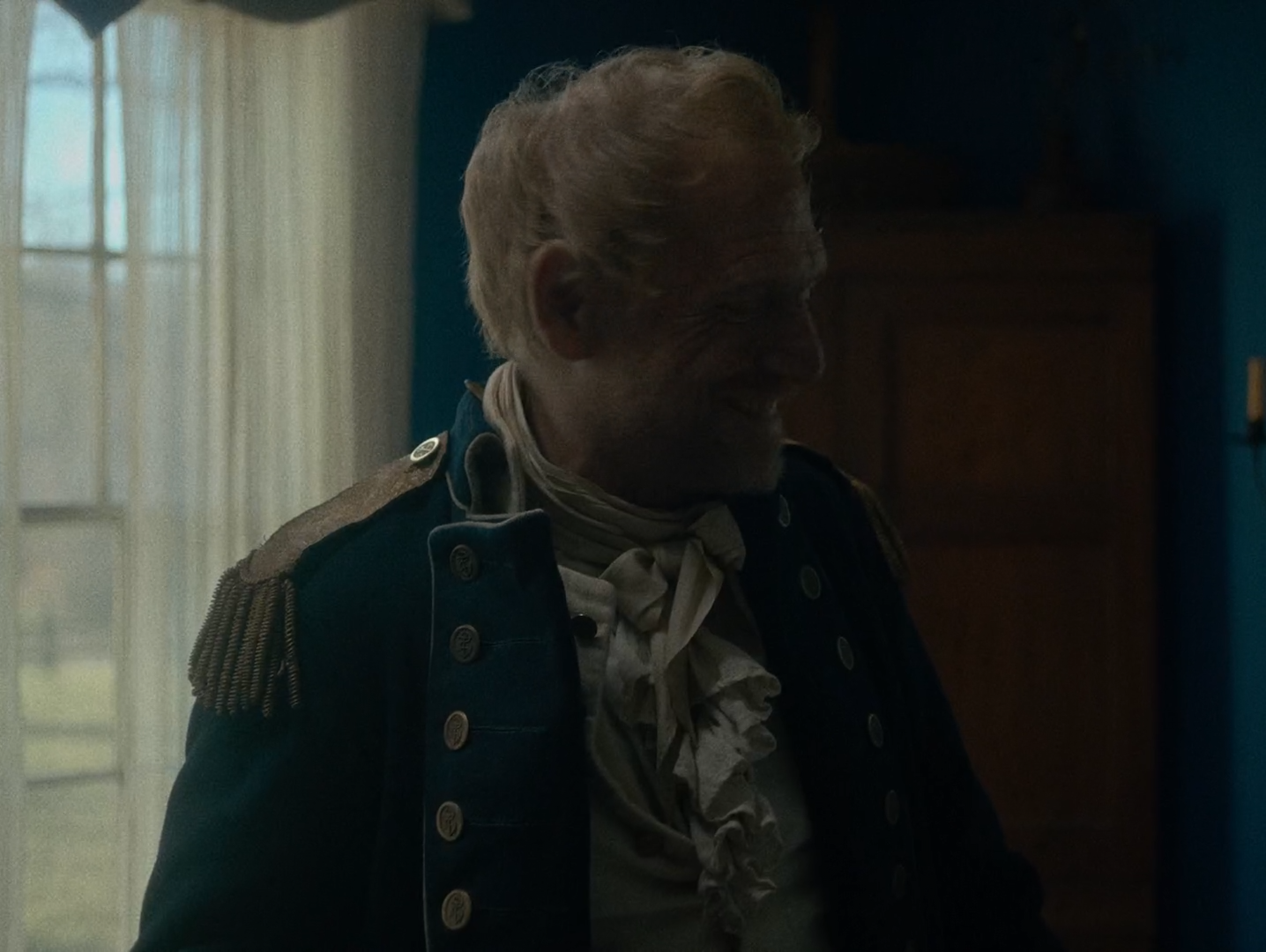 An elderly white-haired man in a dark navy coat with a gold epaulette and white neckcloth sits turned towards a bright curtained window, against a deep blue-green wall.