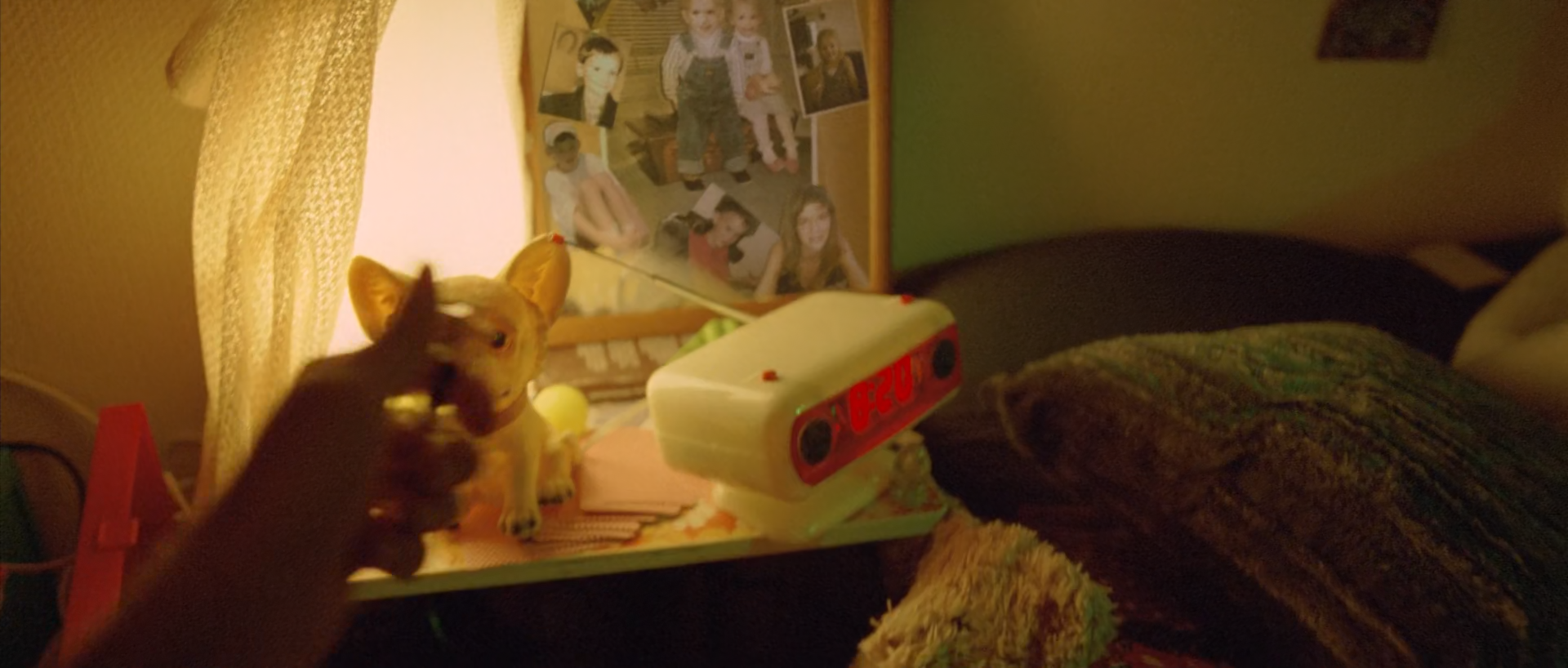 A hand reaches toward two small animal figurines beside a white alarm clock, photographs and a glowing curtain, with a dark bed at right.